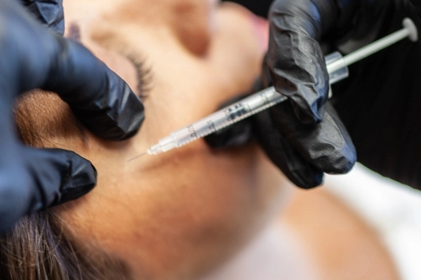 A close-up of a person receiving a skincare treatment, wearing a hairnet, with gloved hands administering a procedure.
